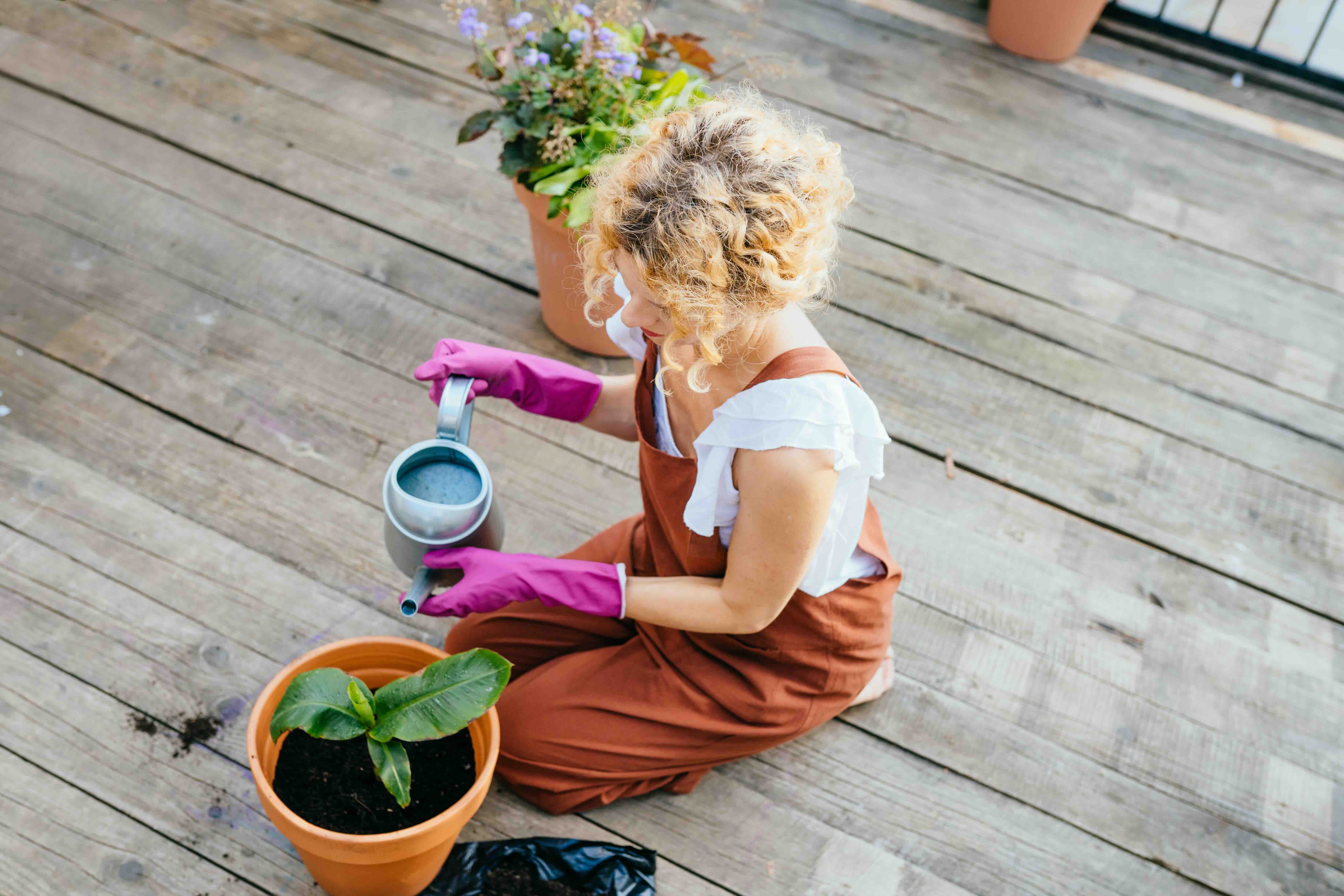 Woman potting and watering a plant on a sunny apartment balcony, surrounded by greenery and urban views.